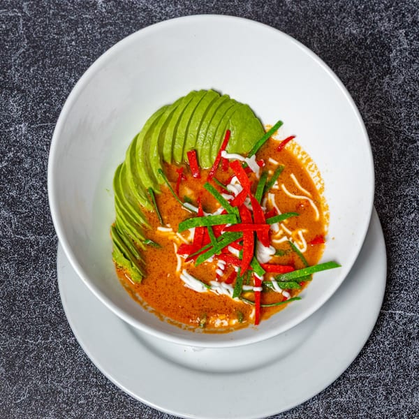 A white bowl of red curry with sliced avocado, red and green peppers, and white sauce.