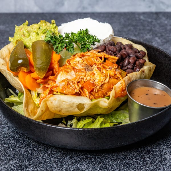A taco salad in a crispy tortilla bowl with chicken, black beans, guacamole, sour cream, and jalapeños.