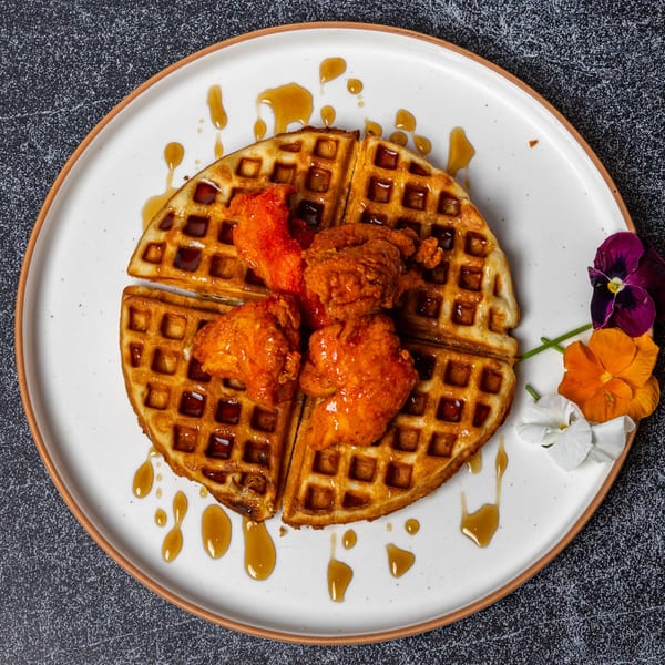 Golden waffle with crispy orange chicken pieces and maple syrup drizzle, garnished with edible flowers.