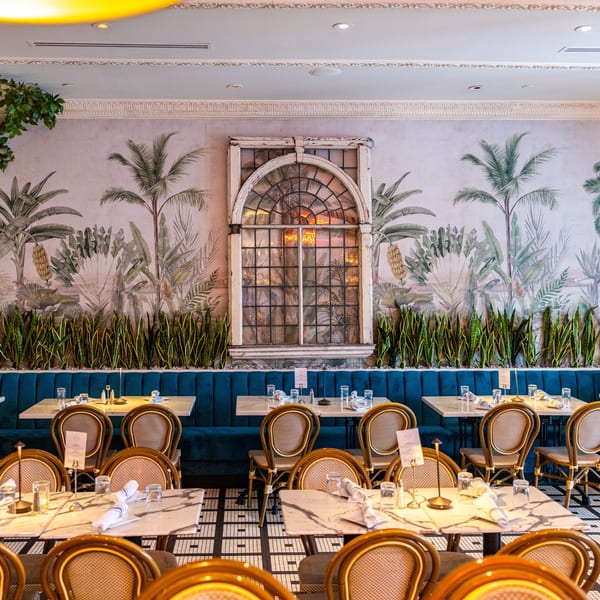 Restaurant interior with blue booths, marble tables, wicker chairs, and a tropical mural with a large window.
