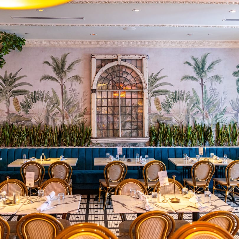 Restaurant interior with blue booths, marble tables, wicker chairs, and a tropical mural with a large window.