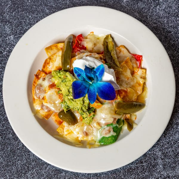 Nachos with melted cheese, guacamole, sour cream, pickled jalapeños, and a blue orchid garnish.