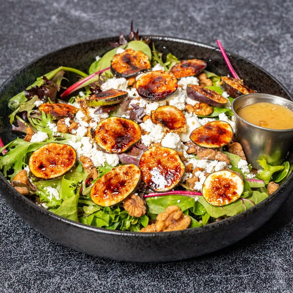 A vibrant salad with mixed greens, caramelized figs, crumbled feta, walnuts, and a side of dressing.