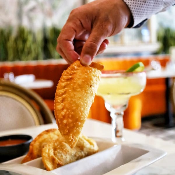 A hand holds a golden-brown empanada over a white dish with more empanadas and a margarita.
