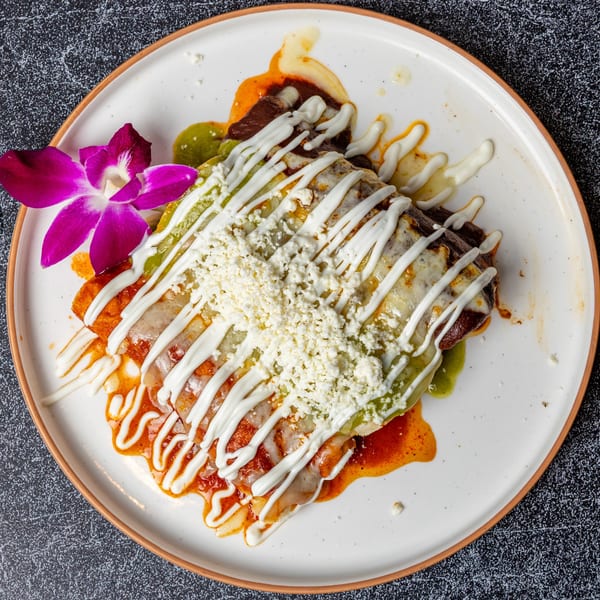 Enchiladas with red and green sauce, drizzled with crema, topped with cotija cheese and a purple orchid.