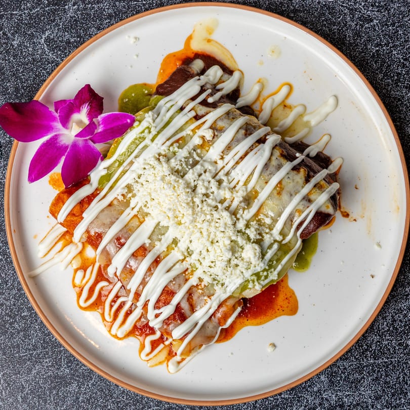 Enchiladas with red and green sauce, drizzled with crema, topped with cotija cheese and a purple orchid.