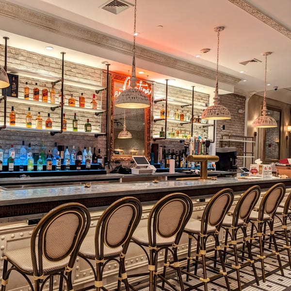 A long, well-lit bar with bottles of liquor on shelves, bar stools, and a neon sign.