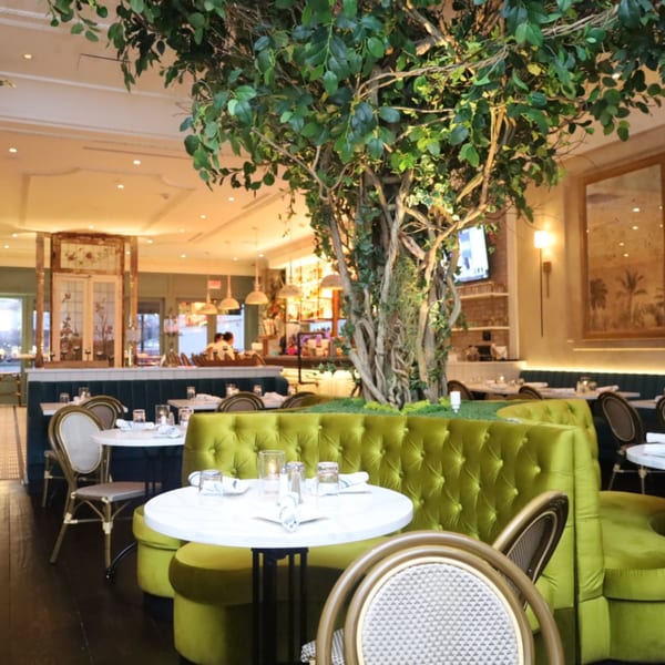 Restaurant interior with green velvet booths, white marble tables, and a large artificial tree in the center.
